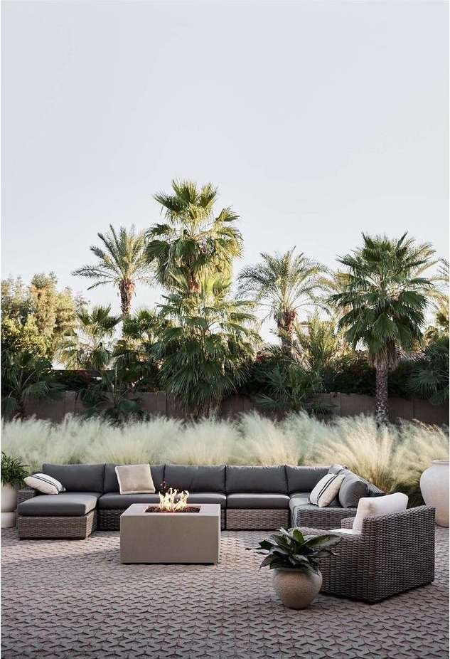Outdoor patio furniture set with a fire pit on a stone patio, surrounded by palm trees and greenery in daylight.