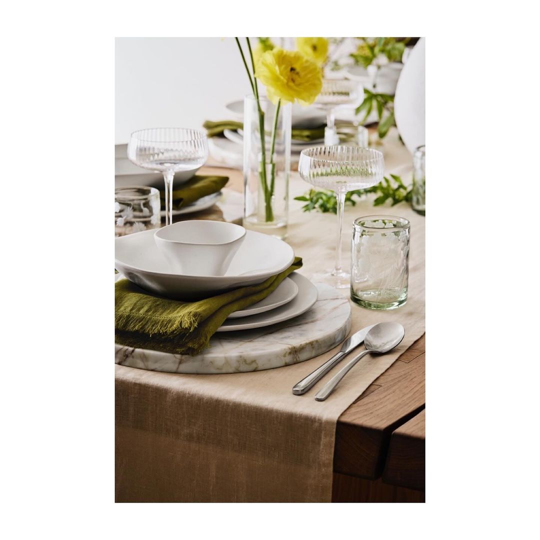 A table setting features white plates, bowls, glassware, and silverware on a beige runner with a marble board and yellow flowers.