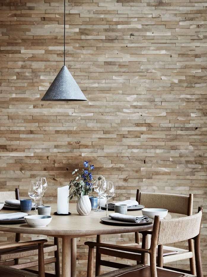 A modern dining room setup featuring a round wooden table with place settings, chairs, and a cone-shaped pendant light against a brick wall.