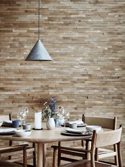 A modern dining room setup featuring a round wooden table with place settings, chairs, and a cone-shaped pendant light against a brick wall.