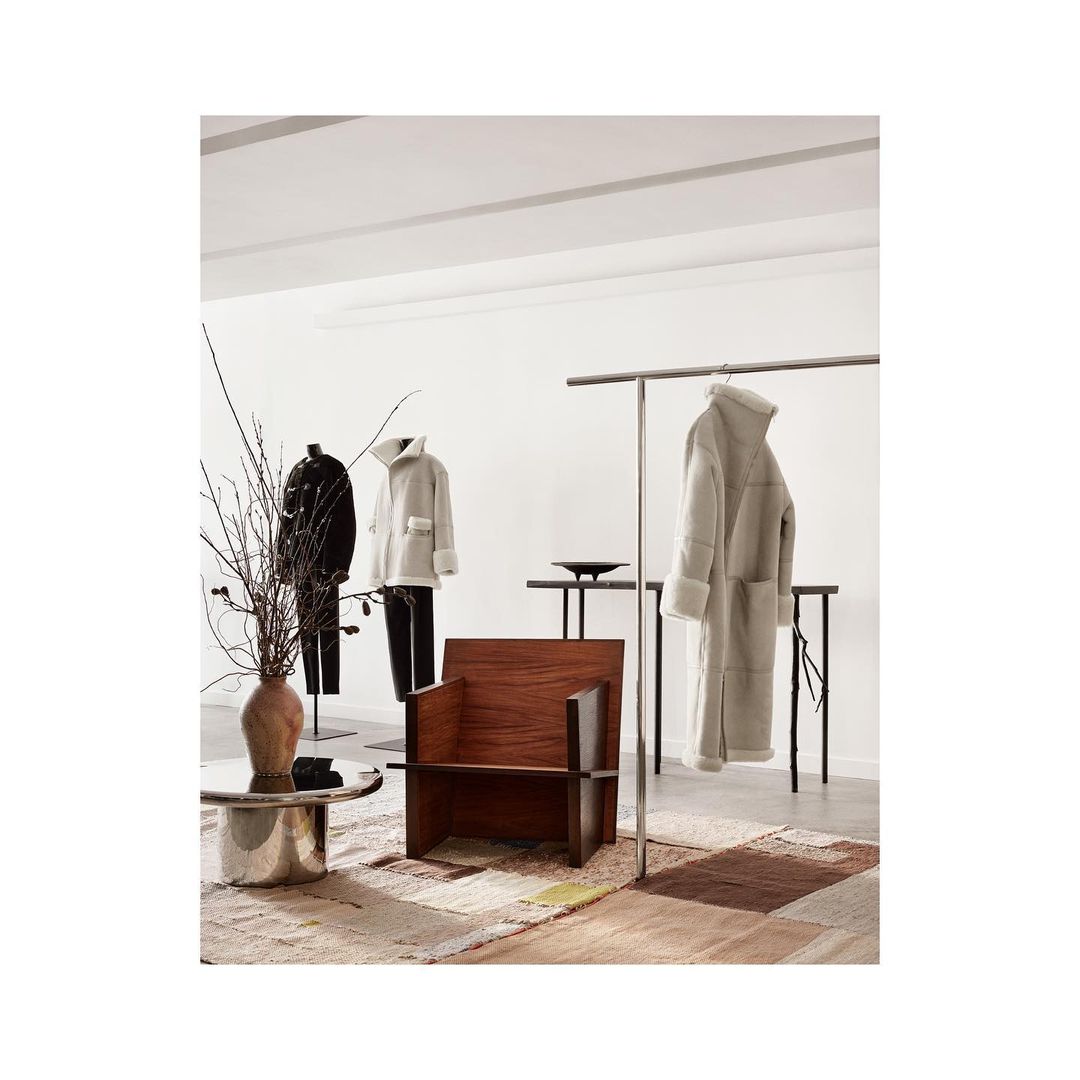 Interior of a retail space featuring a wooden chair, metal coffee table, and clothing displayed on racks and mannequins against a white backdrop.