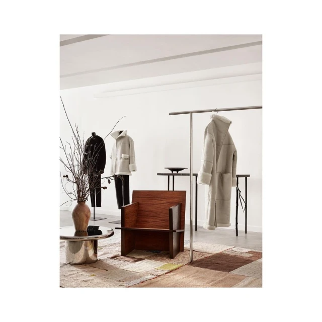 Interior of a retail space featuring a wooden chair, metal coffee table, and clothing displayed on racks and mannequins against a white backdrop.