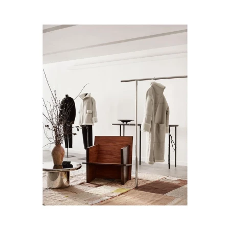 Interior of a retail space featuring a wooden chair, metal coffee table, and clothing displayed on racks and mannequins against a white backdrop.