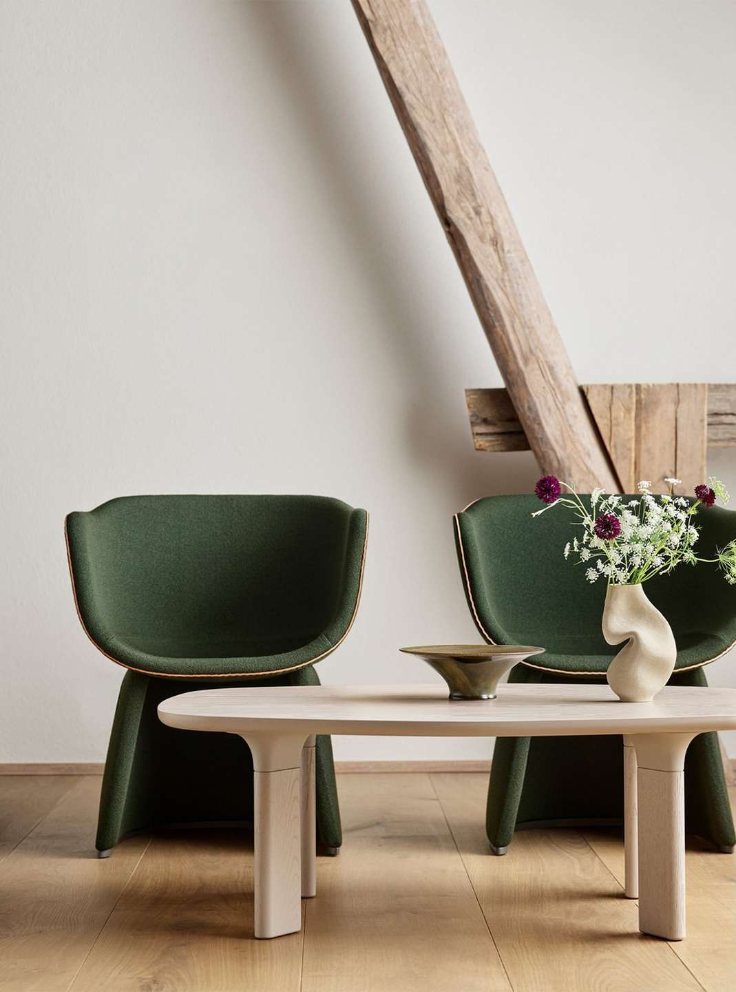 Interior featuring two modern green upholstered armchairs, a light wood coffee table with a bowl, and a vase of flowers in a bright room.