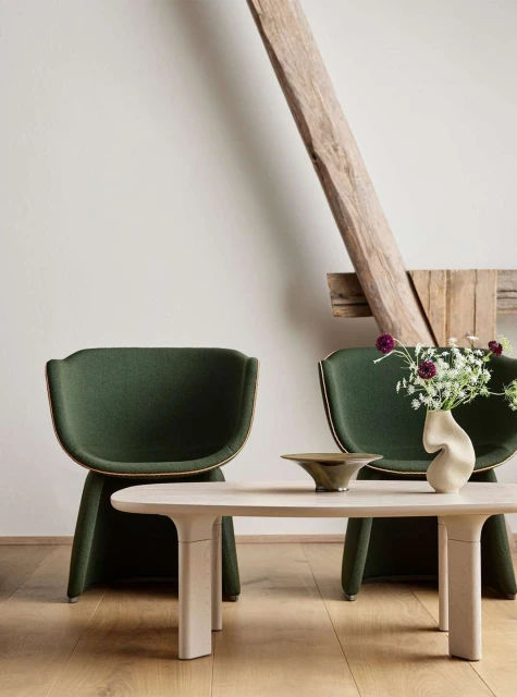 Interior featuring two modern green upholstered armchairs, a light wood coffee table with a bowl, and a vase of flowers in a bright room.