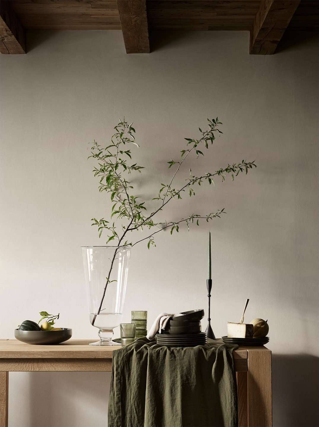 Neutral still life with a clear glass vase holding green branches, surrounded by tableware on a light wood table against a cream wall.