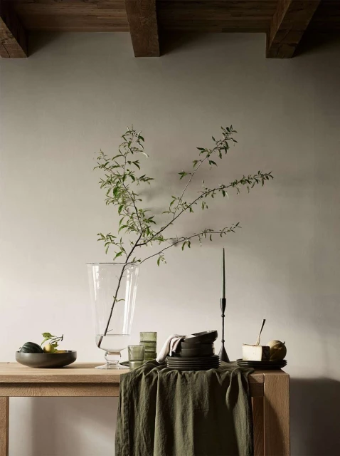 Neutral still life with a clear glass vase holding green branches, surrounded by tableware on a light wood table against a cream wall.