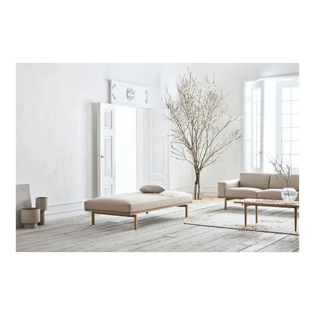 Beige sofa and daybed with throw pillow in a well-lit, minimalist interior with white walls, light wood floors, and a decorative tree.