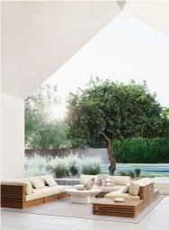 Outdoor patio lounge area with wooden sectional sofas and coffee table under a large tree against a white modern architectural overhang.