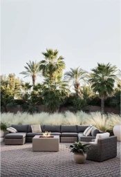 Outdoor patio lounge set with grey sectional sofa and fire pit table against a backdrop of palm trees and tall ornamental grass.