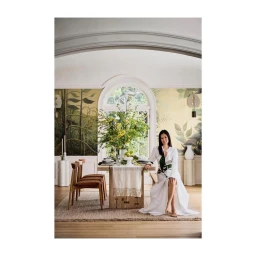 Woman in white dress seated at a wooden dining table in a room with botanical wallpaper, photographed by Anders Schonnemann.