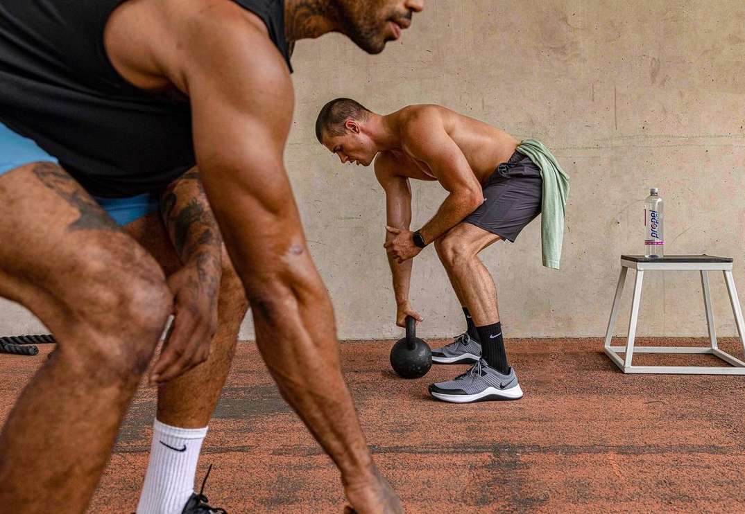 Two muscular men engage in an intense kettlebell workout at an indoor gym with a water bottle on a stool nearby.