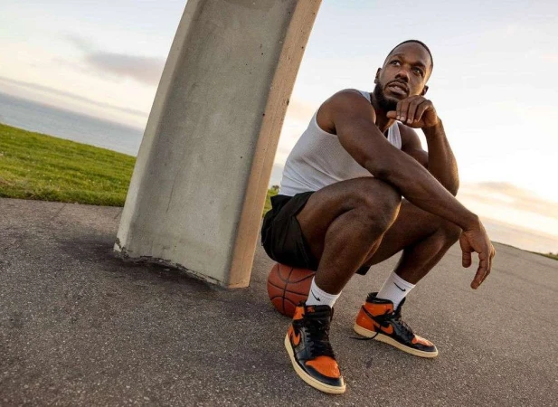 Black man crouching outdoors near a concrete pillar resting his chin on his hand while perched on a basketball