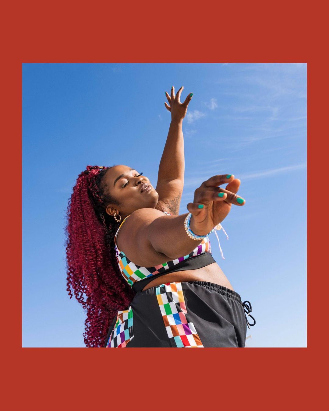 Woman with red curly hair in a color-block workout outfit stretches her arms toward the blue sky in a low-angle outdoor shot.