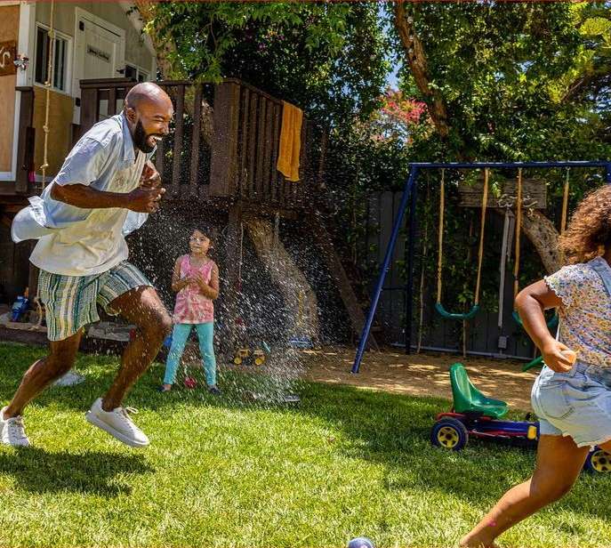 A man and a girl are running through a sprinkler in a sunlit backyard, with a second girl standing nearby. Trees and playground equipment fill the background.