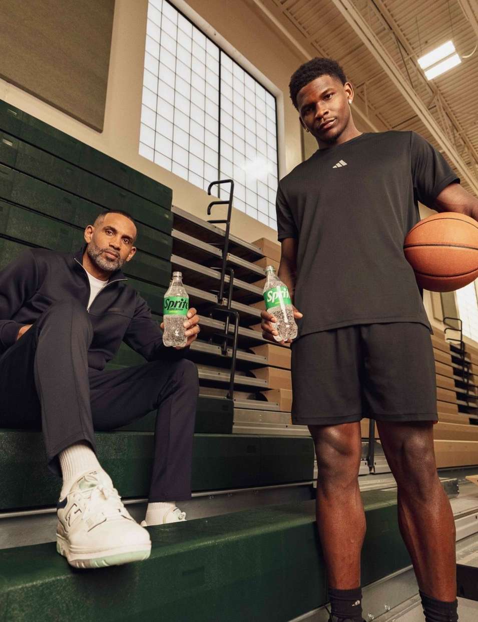 Two African-American men seated on green bleachers indoors, one holding a basketball, both holding Sprite bottles, natural lighting.