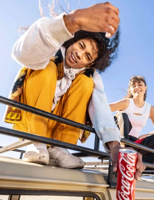 Two young adults pose with Coca-Cola on top of a safari vehicle on a sunny day with a clear blue sky, showcasing youthful energy.