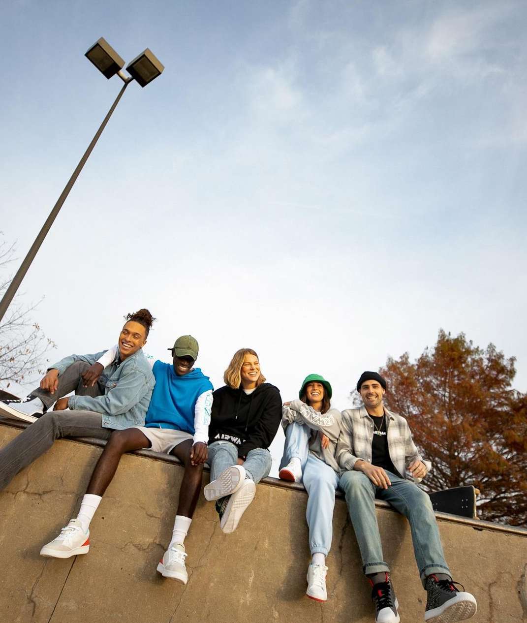 Group of five young adults in casual clothing sitting outdoors at a skate park on a sunny day with a clear blue sky.