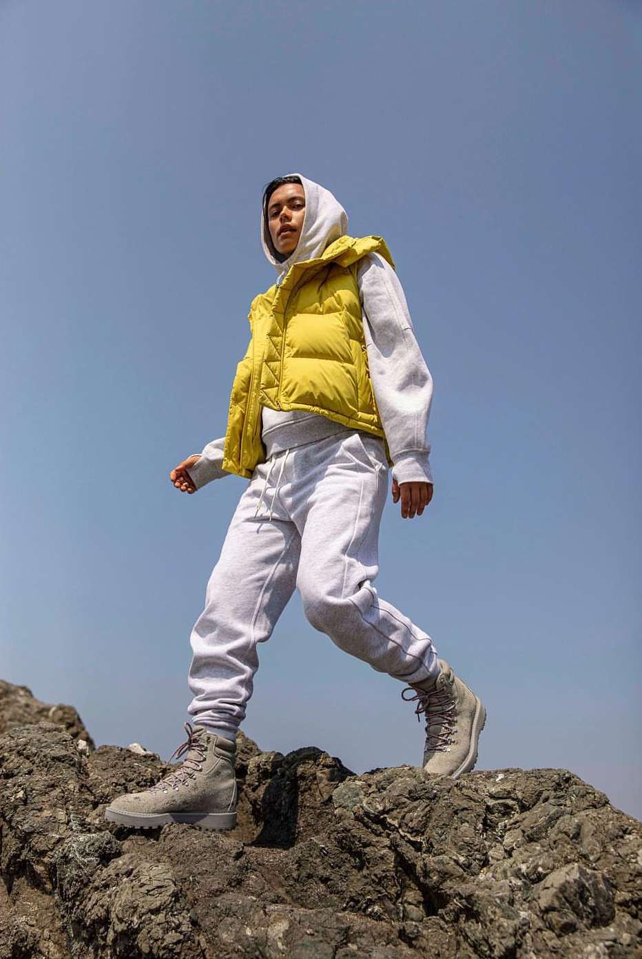 Adult wearing a yellow puffer vest and gray sweats walking on rocks against a blue sky. Boots with laces are visible.