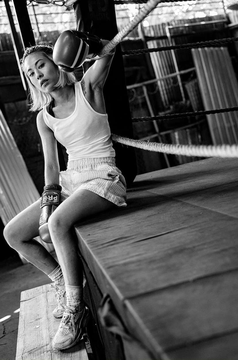 Black and white portrait of an Asian female boxer resting in the corner of a boxing ring wearing boxing gloves, shorts, and a tank top.