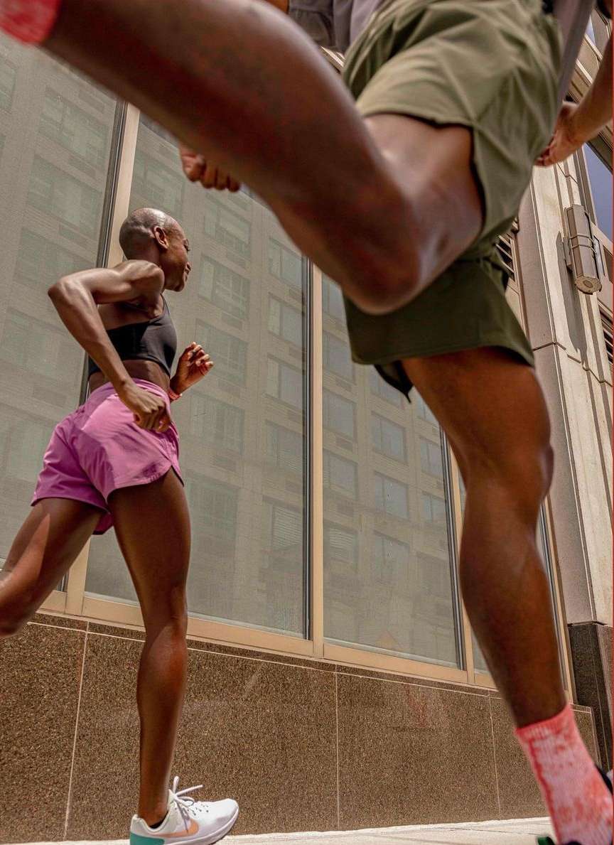 Two athletic adults are running outdoors in pink and green athletic shorts and matching shoes, with a city building in the background.
