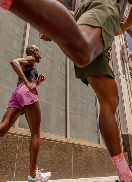 Two athletic adults are running outdoors in pink and green athletic shorts and matching shoes, with a city building in the background.