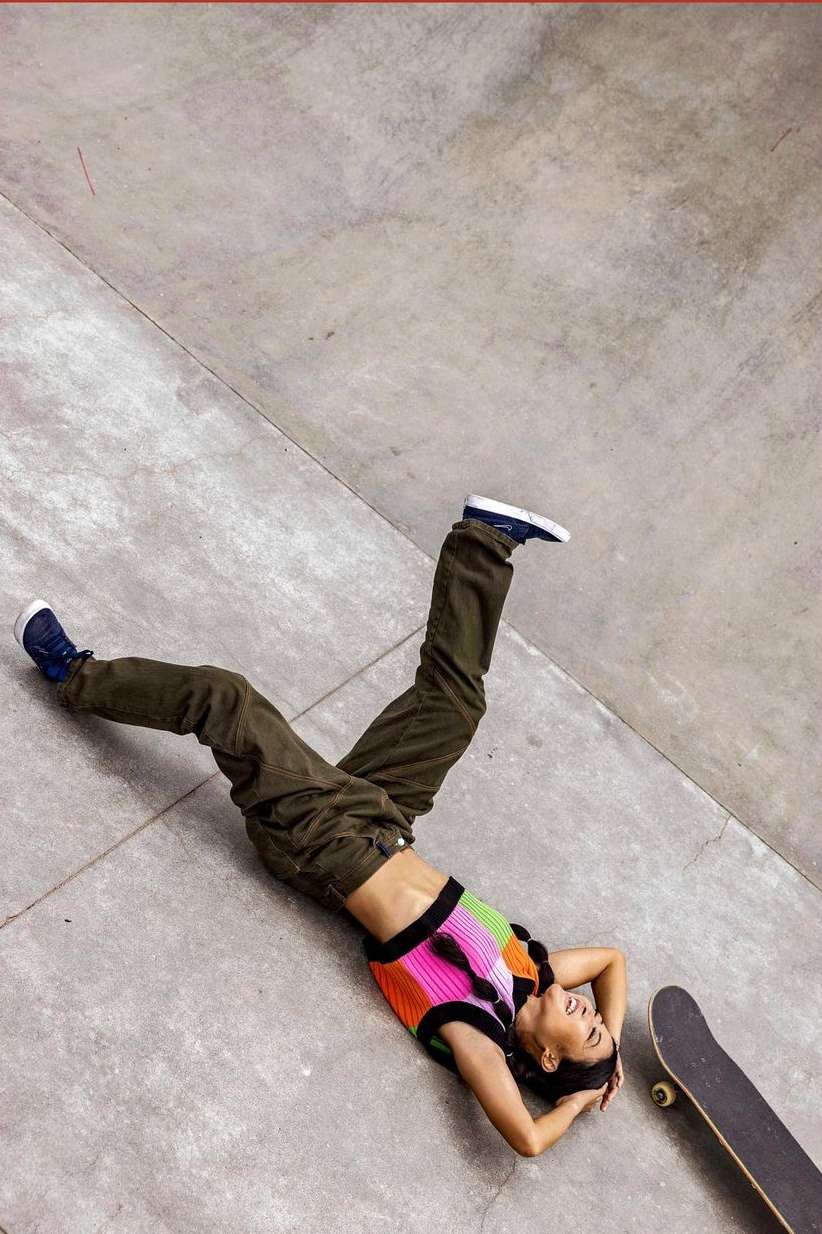 Woman with skateboard laying on grey concrete with an overhead view, wearing a colorful crop top and dark pants, looking up.