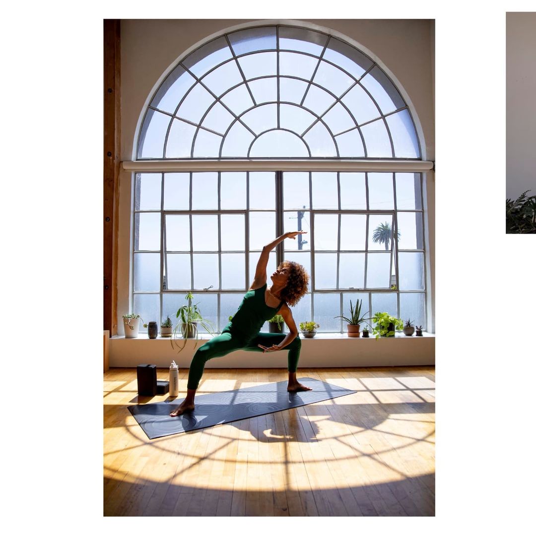 Woman practices yoga in green athletic wear on a mat in a sunlit room with large arched window and plants in a fitness lifestyle scene.