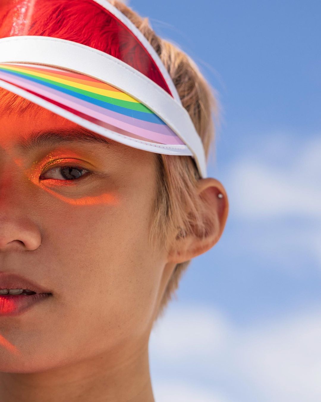 Close-up portrait of a young adult with rainbow visor, shimmery eyeshadow, fair skin, and short blonde hair against a light blue sky.