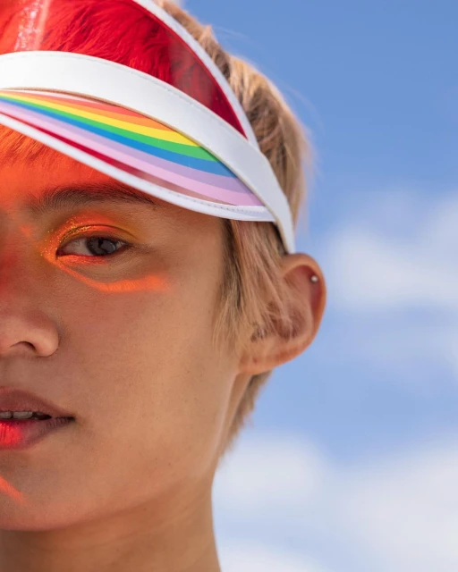 Close-up portrait of a young adult with rainbow visor, shimmery eyeshadow, fair skin, and short blonde hair against a light blue sky.