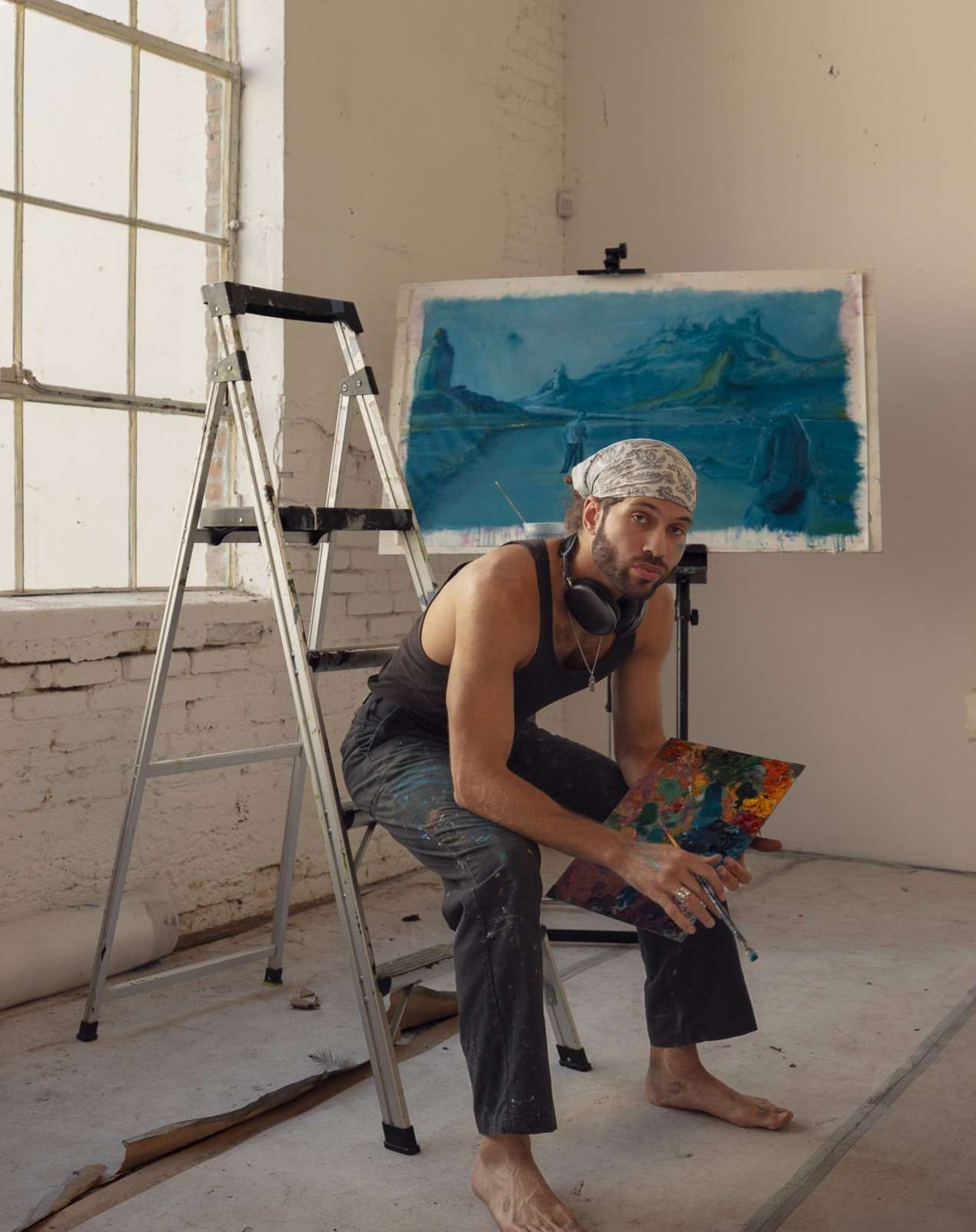 Painter in tank top sits on ladder in studio with his painting. The artist wears headphones and holds his color palette.