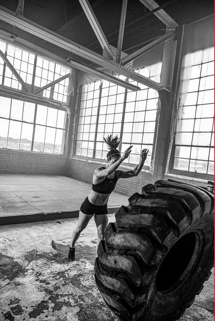 A woman in athletic wear is working out inside a rustic brick building, pushing a large tire with natural lighting from window in daytime.