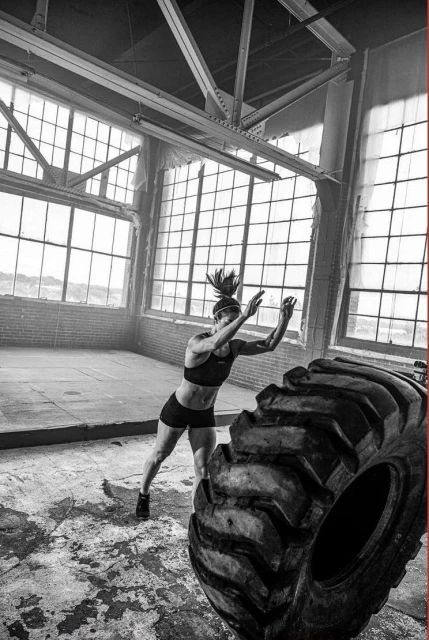 A woman in athletic wear is working out inside a rustic brick building, pushing a large tire with natural lighting from window in daytime.