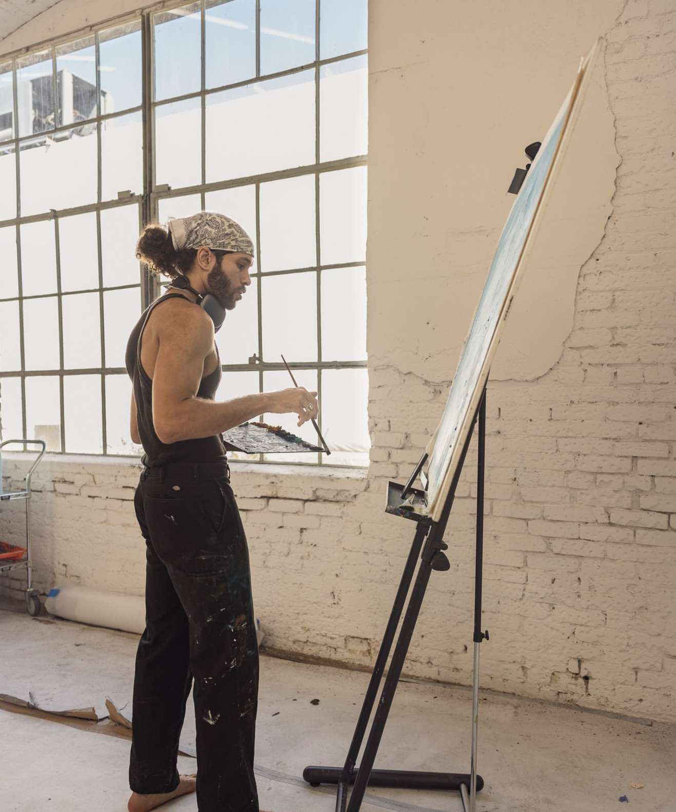 Man with bandana stands indoors painting on a canvas in a spacious, naturally lit studio with paint-splattered clothing and bare feet.