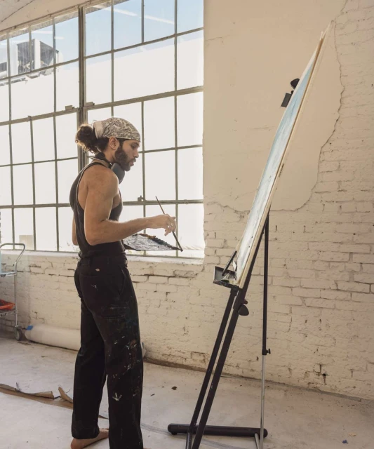Man with bandana stands indoors painting on a canvas in a spacious, naturally lit studio with paint-splattered clothing and bare feet.