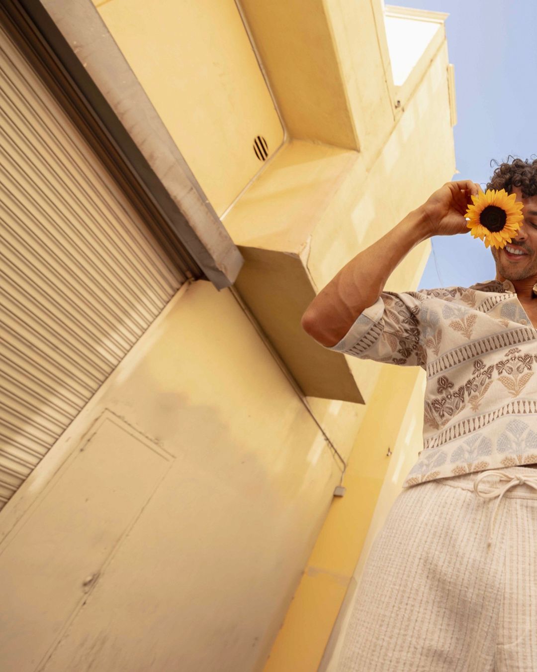 A smiling adult holds a sunflower to their eye, set against a yellow building and clear blue sky. Low-angle, eye-level shot.