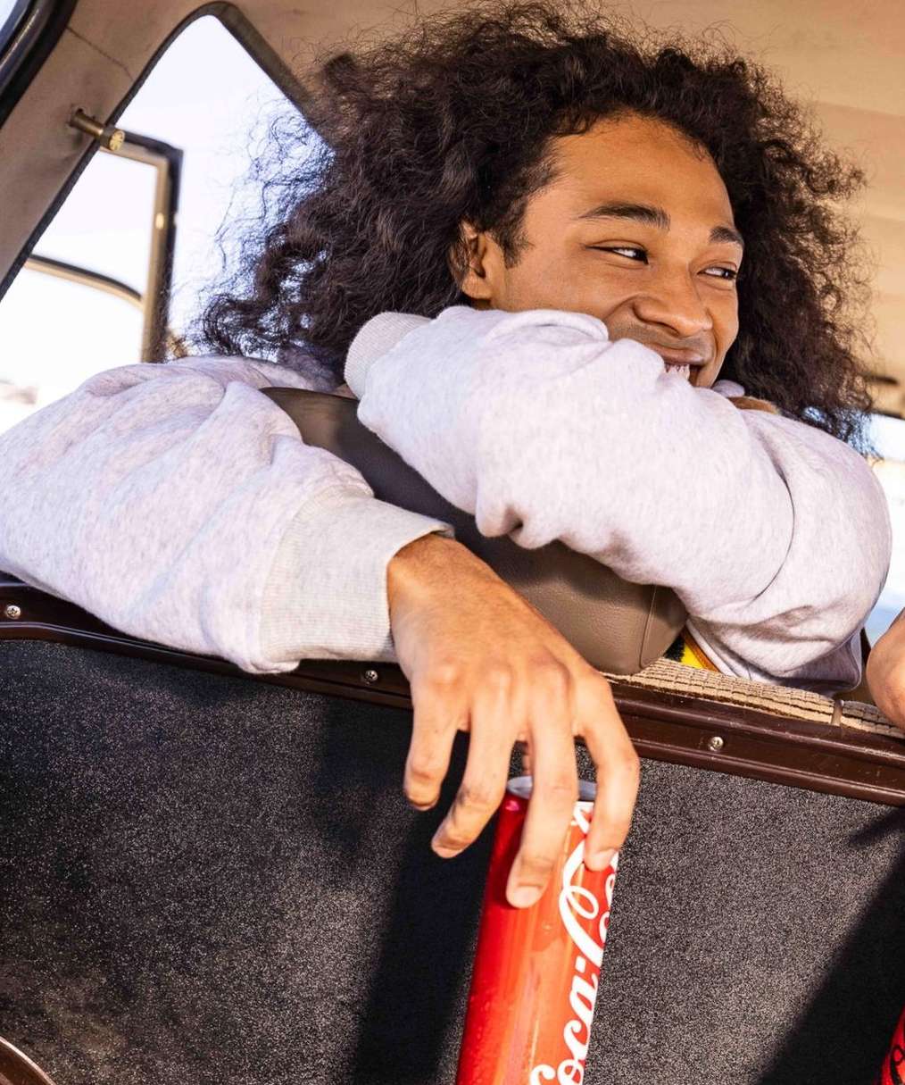 Man with curly dark hair smiles holding a red Coca-Cola can inside of a car looking out the open window on a sunny day.