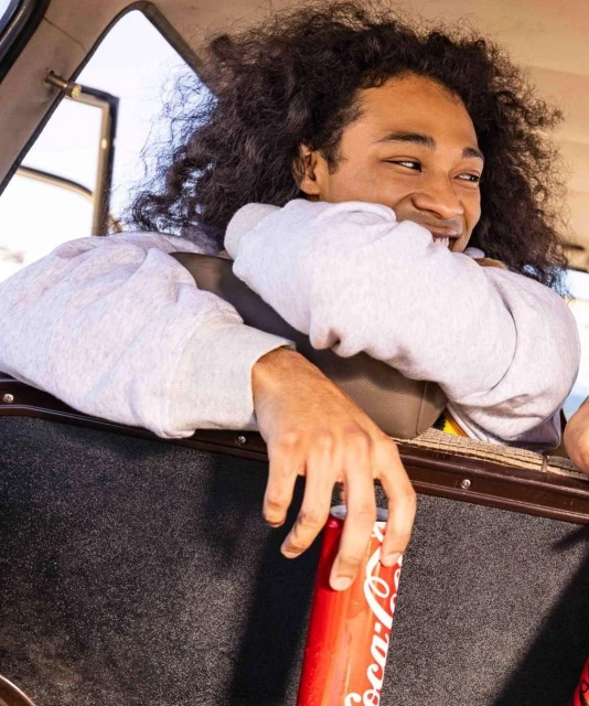 Man with curly dark hair smiles holding a red Coca-Cola can inside of a car looking out the open window on a sunny day.