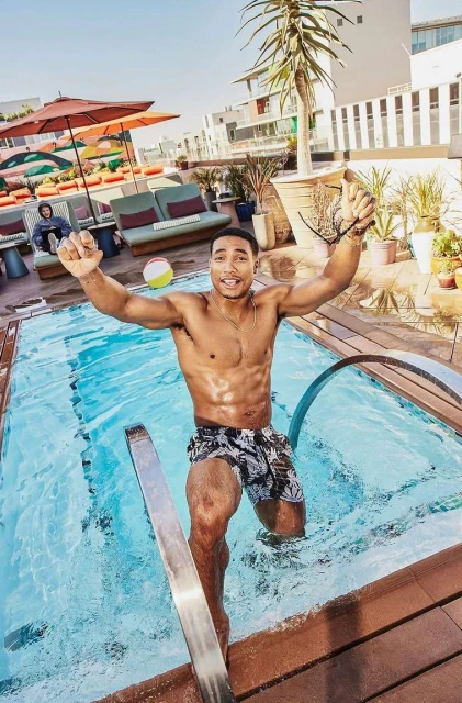 Fit man with arms raised exiting an outdoor swimming pool at a rooftop patio area on a sunny day wearing patterned swimming trunks.