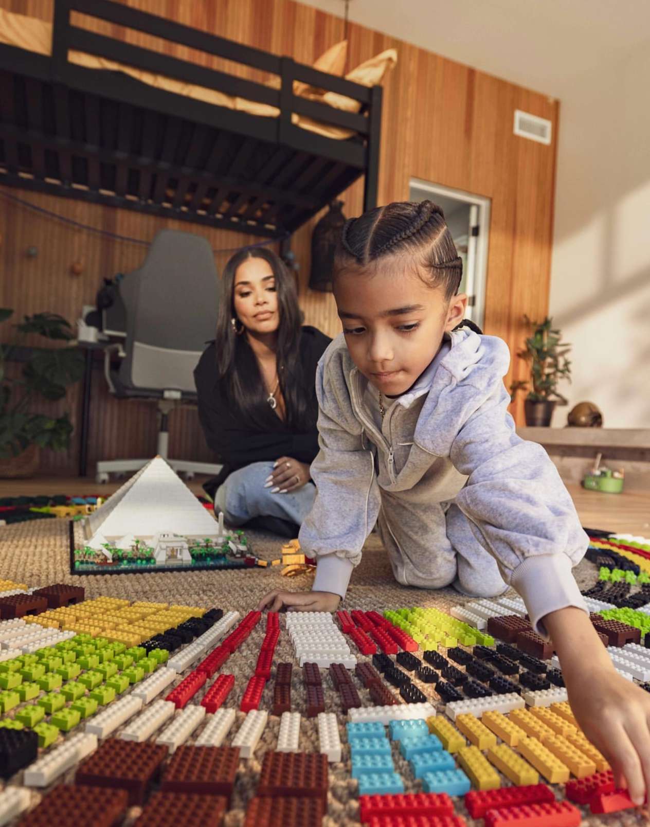 Child and adult woman play with toy building blocks inside a wood-paneled room with bunk beds and a carpeted floor, daytime setting.