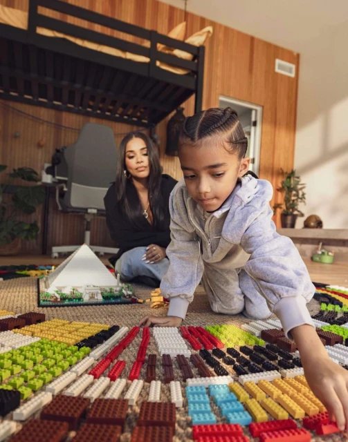 Child and adult woman play with toy building blocks inside a wood-paneled room with bunk beds and a carpeted floor, daytime setting.