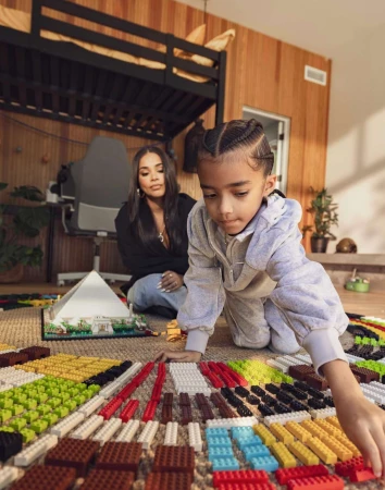 Child and adult woman play with toy building blocks inside a wood-paneled room with bunk beds and a carpeted floor, daytime setting.