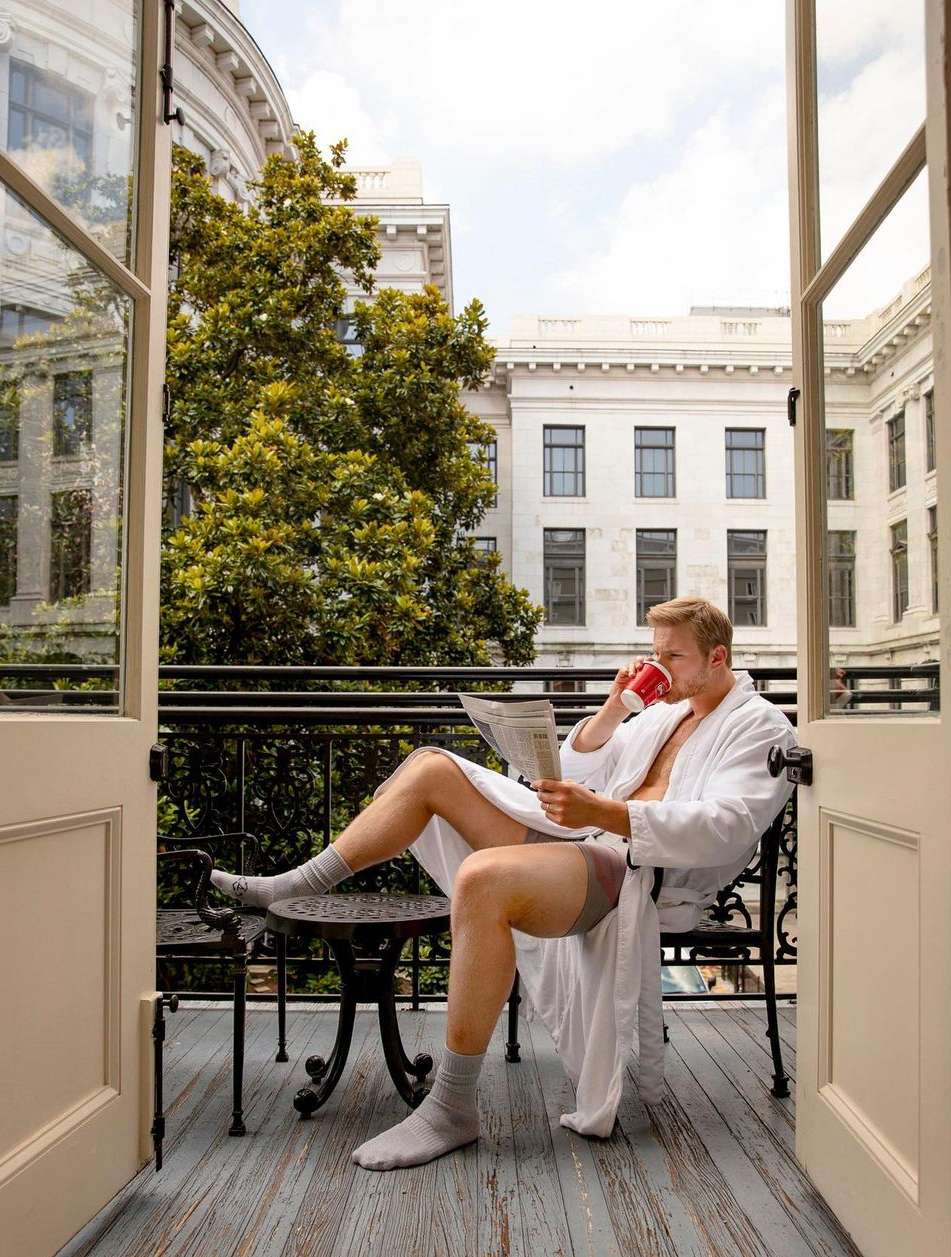Man in bathrobe sits on balcony reading newspaper and drinking coffee. Exterior of white building with many windows in background.