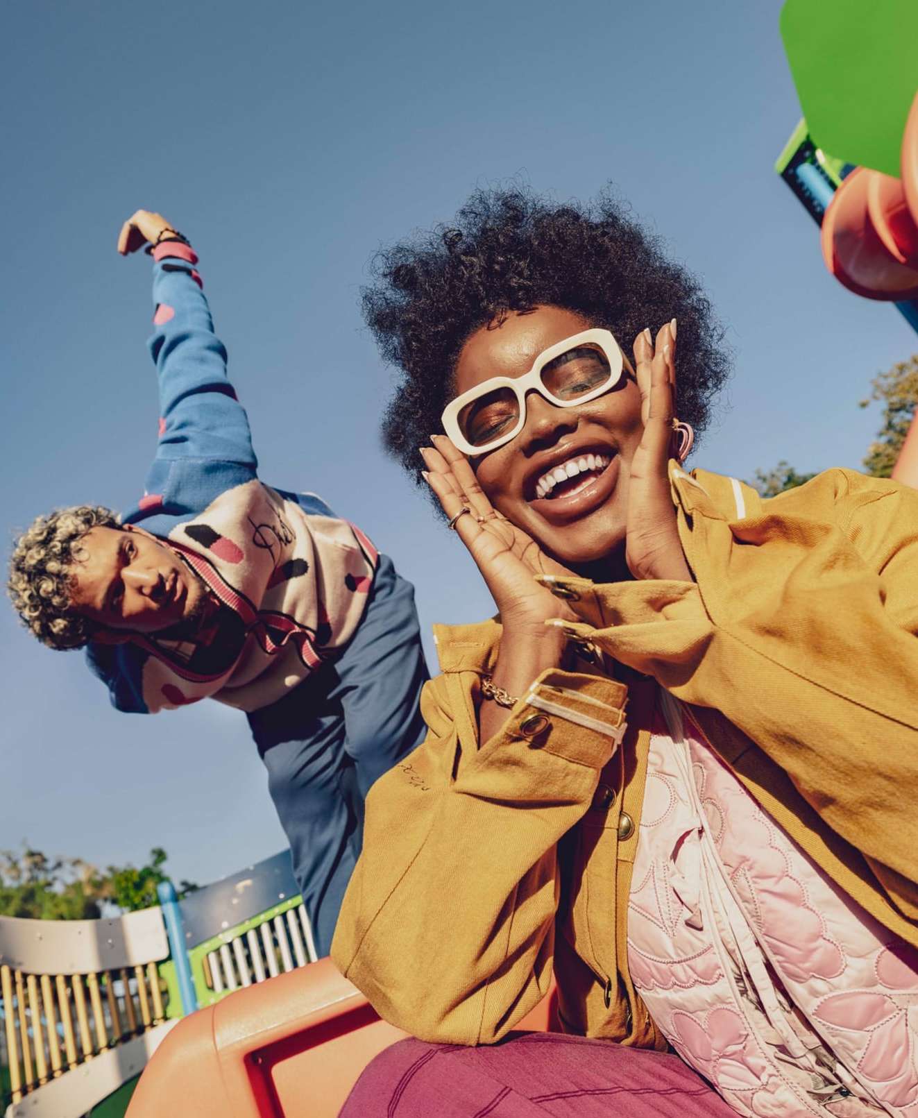 Two friends smiling at the camera while enjoying an outdoor playground on a sunny day. A cheerful lifestyle scene with a low-angle perspective.