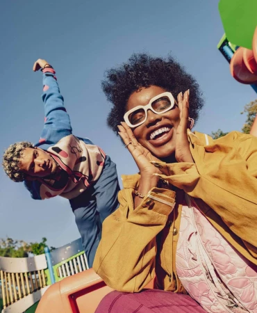Two friends smiling at the camera while enjoying an outdoor playground on a sunny day. A cheerful lifestyle scene with a low-angle perspective.