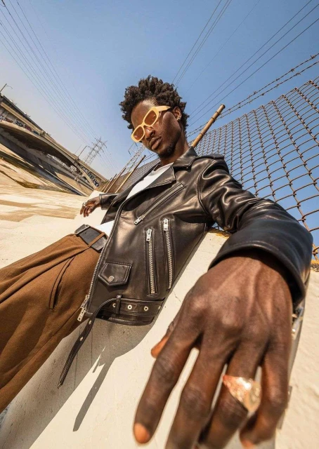 Stylish man poses in a leather jacket and sunglasses in an urban environment with bridge and fences on a sunny day.