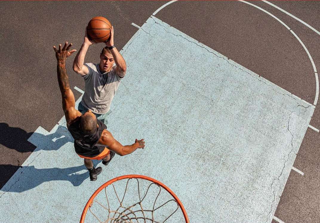 Overhead shot of two adult men playing basketball on an outdoor court, one shooting and one defending, under bright daylight.