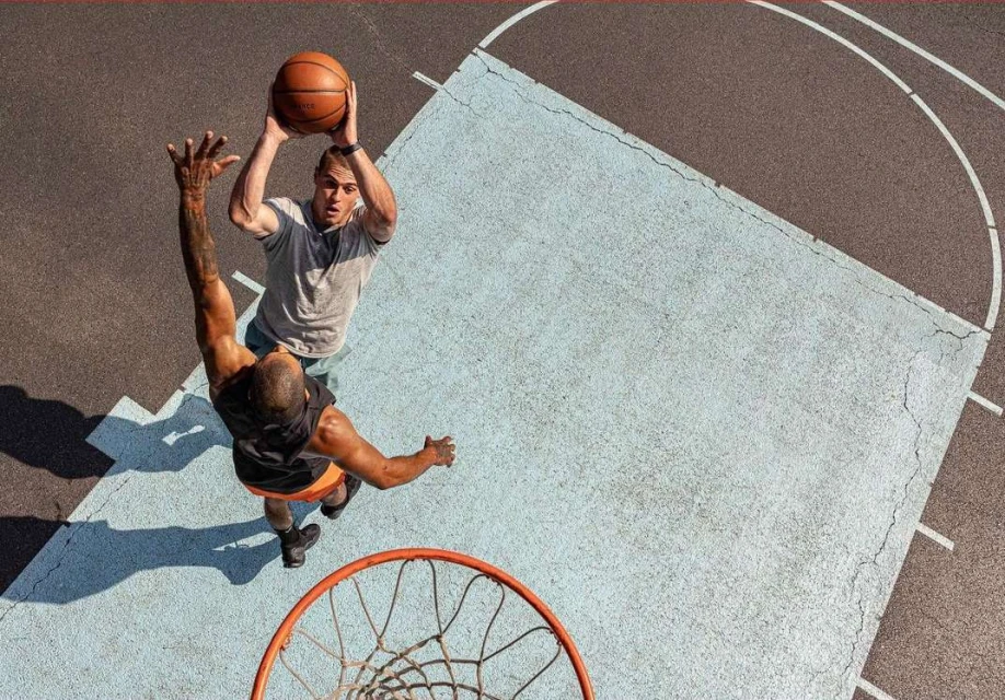 Overhead shot of two adult men playing basketball on an outdoor court, one shooting and one defending, under bright daylight.