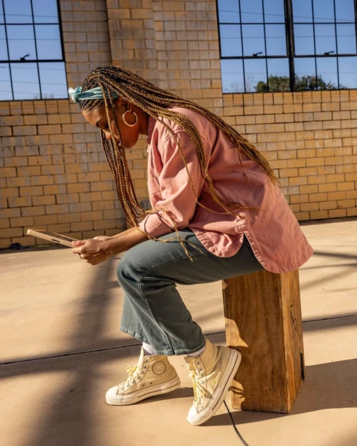 Stylish woman with braided hair sits on a wooden box while looking at a tablet outdoors on concrete in the sunlight.