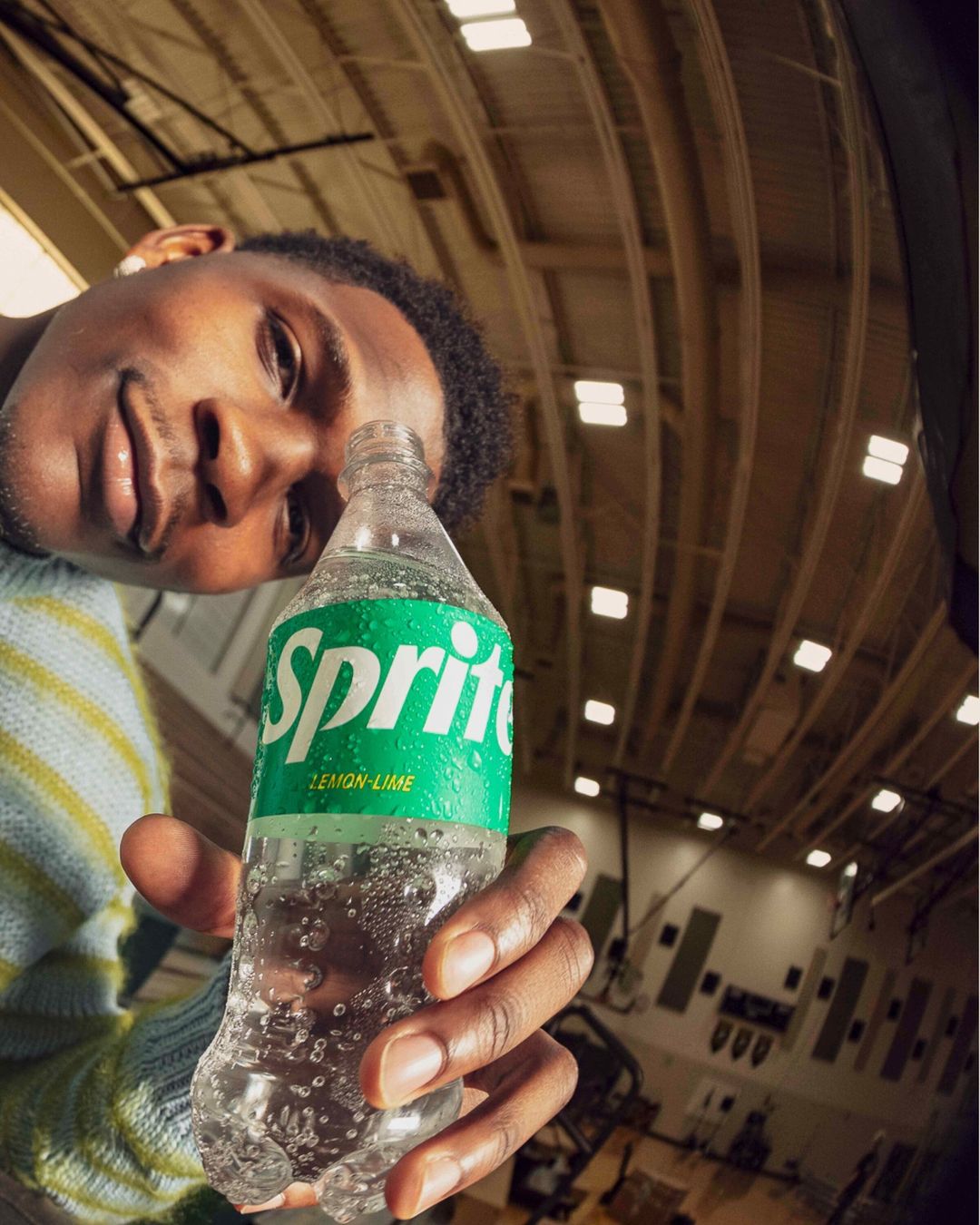 Man holding a Sprite bottle in an indoor sports arena. The fisheye-lens image focuses on the bottle's label and cap, with the man's face.